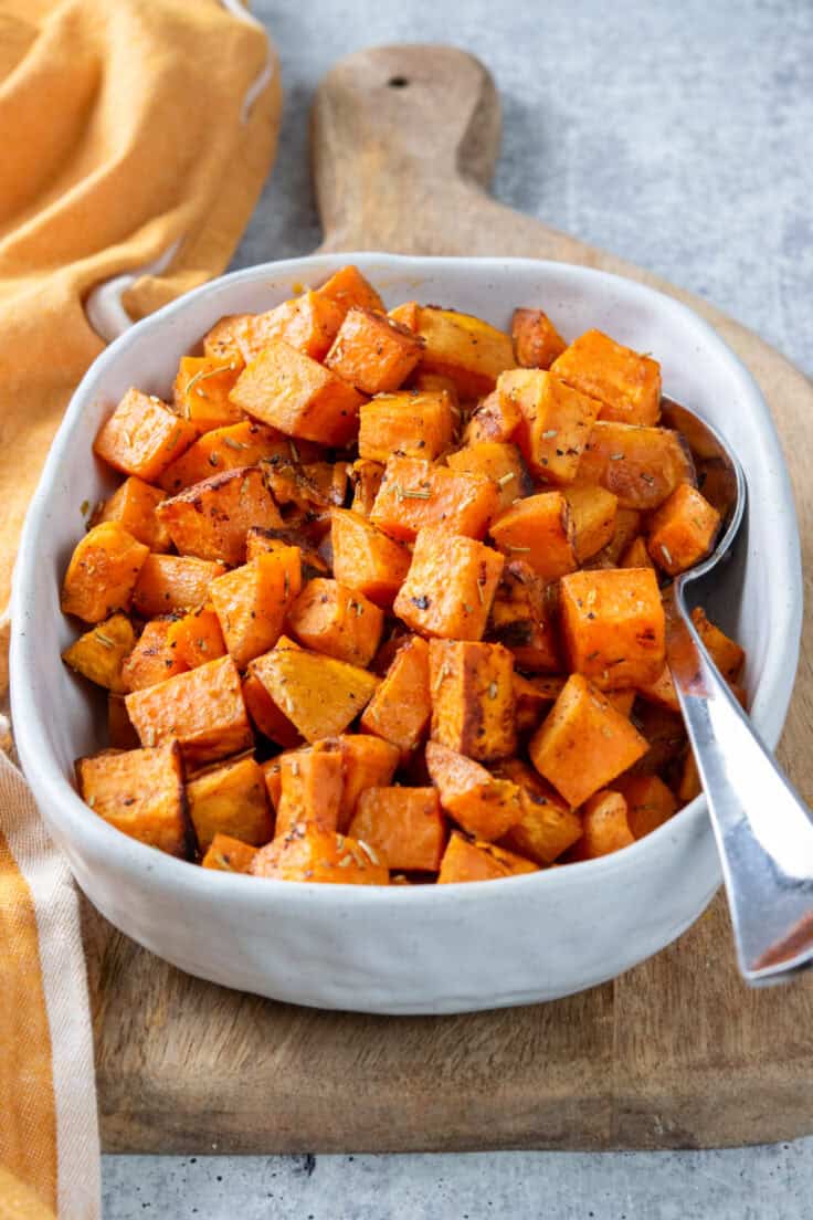 A serving dish of roasted sweet potatoes. You can see roasted bits and caramelization and the seasoning on the potatoes. There's a serving spoon in the dish.