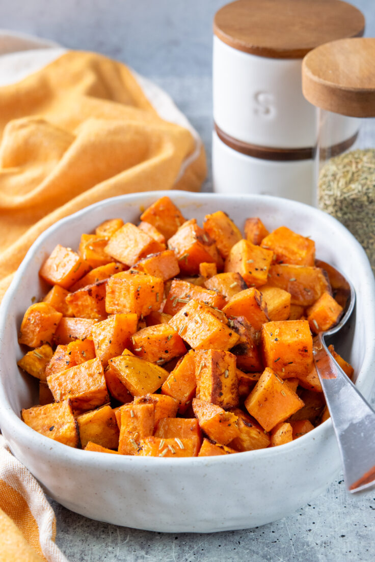 Roasted sweet potatoes in a white oval serving dish next to a rosemary spice jar and salt and pepper.