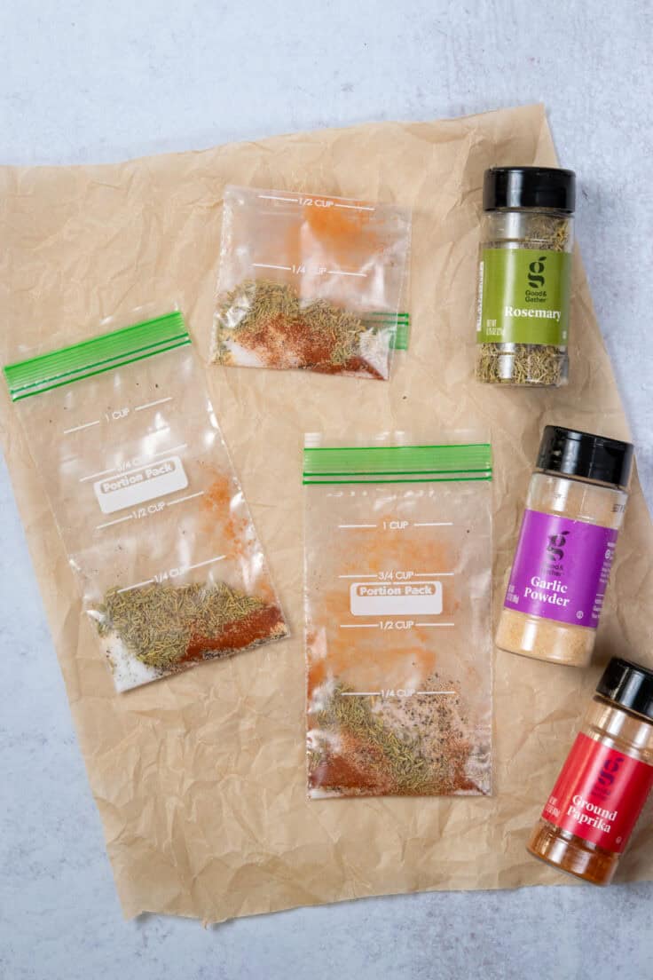 Three homemade seasoning packets in baggies for making roasted sweet potatoes. There are jars of rosemary, garlic powder and paprika next to the packets.