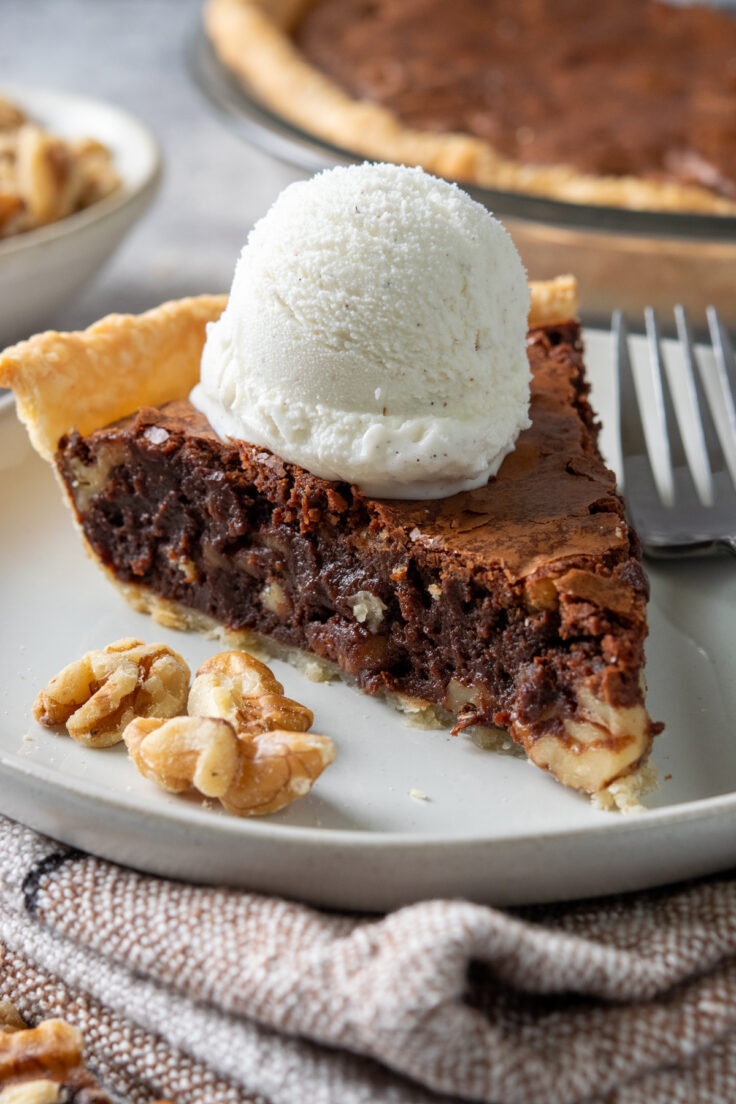 A delicious slice of fudge pie on a dessert plate with a fork. The chocolate pie is served a la mode with vanilla ice cream. You can see gooey bits of chocolate and walnuts in the pie.