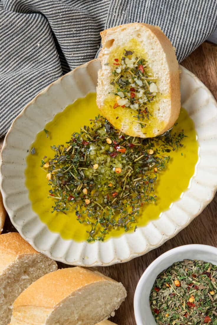 A slice of Italian bread in a dish of bread dipping oil. The dish is surrounded by thick slices of bread for dipping and a small bowl with the seasoning blend for the olive oil.