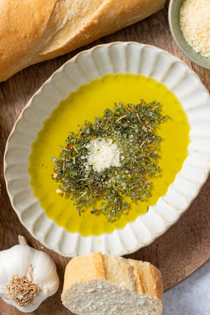 A bowl of bread dipping oil on a tray with bread. There's a buld of garlic and small bowl of grated parmesan next to the dipping sauce. The cheese has just been added on top of the seasoning mix in the oil.