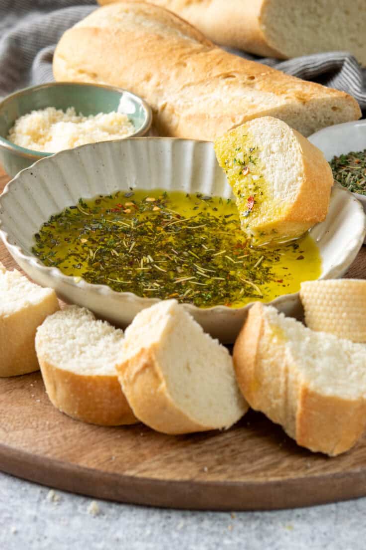 Bread dipping oil served on a tray with thick slices of bread cut in half so there's a side without crust so the bread soaks up the olive oil and herbs. Next to the tray is a loaf of the crusty bread and a small bowl of parmesan cheese.