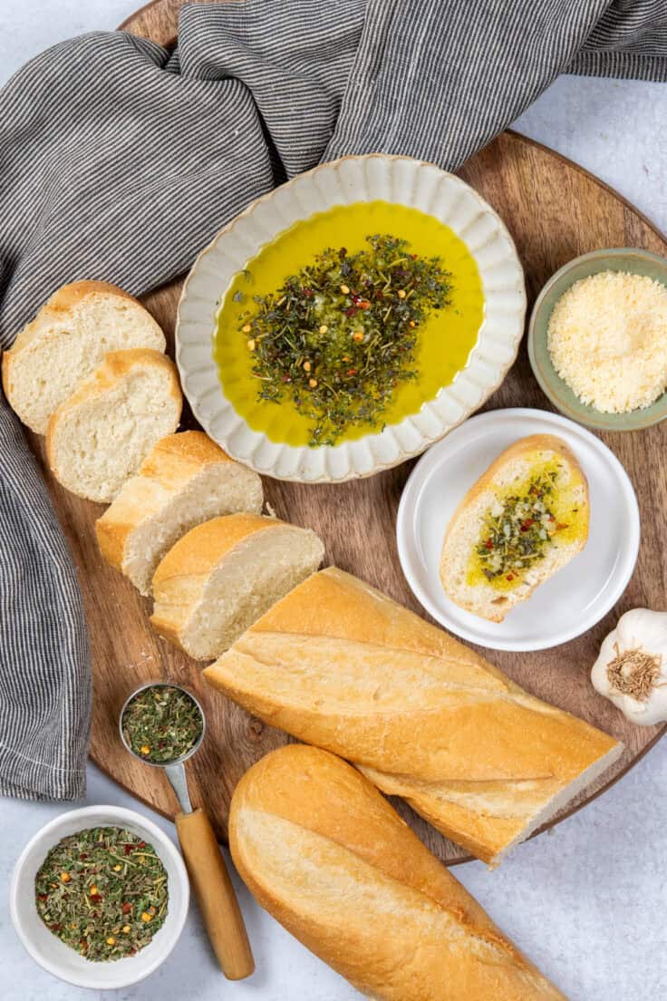 An overhead view showing the bread dipping sauce in a shallow dish, a long loaf of French bread cut into thick slices, the Italian seasoning blend for adding to the oil, as well as a bulb of garlic and bowl of grated Parmesan. There's also a piece of bread covered in the olive oil dip.
