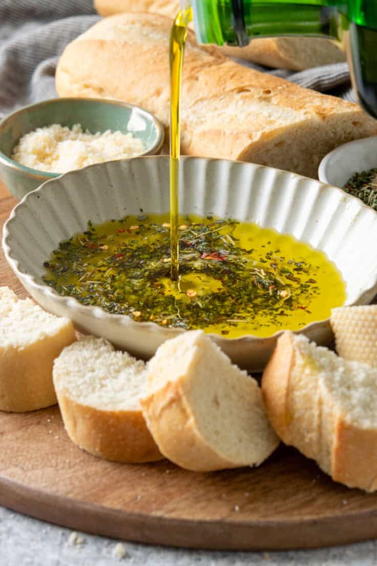 Pouring olive oil into a dish with Italian seasoning blend to make the dipping oil. The dish is on a wood tray that has sliced French bread on it and a small bowl of parmesan cheese.