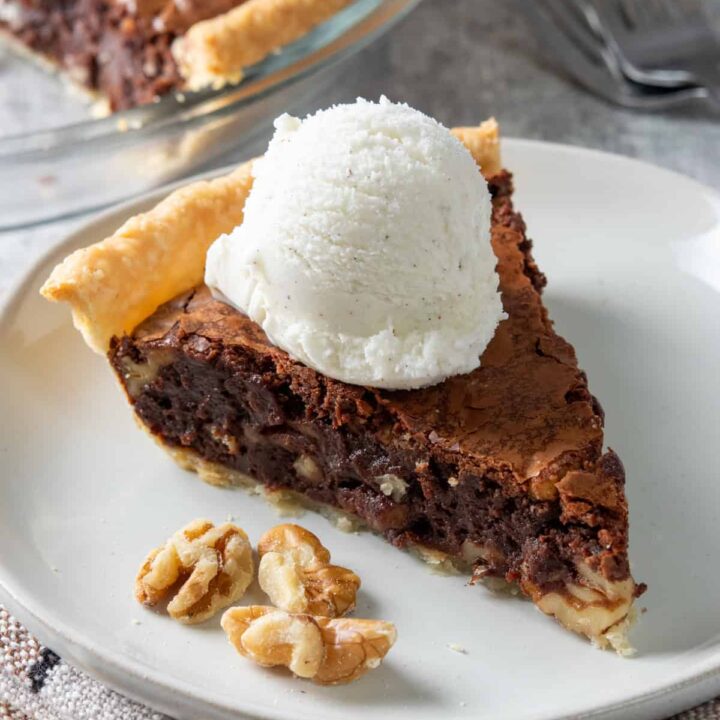 A gooey slice of chocolate fudge pie with ice cream on top. It has walnuts in it and is on a plate next to the pie plate.