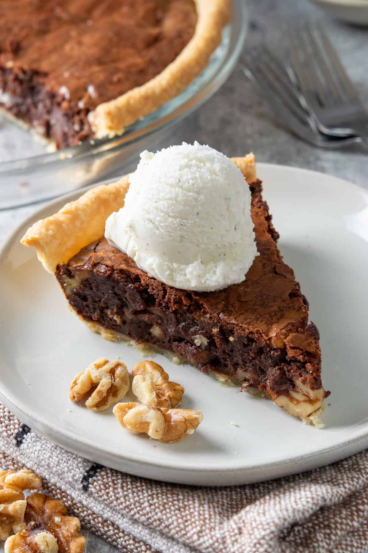 A gooey slice of chocolate fudge pie with ice cream on top. It has walnuts in it and is on a plate next to the pie plate.
