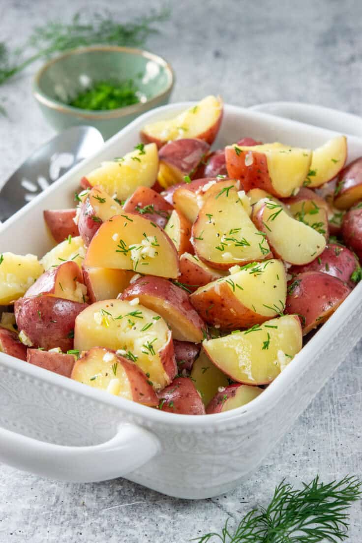 Garlic dill new potatoes in a serving dish with a serving spoon next to it. The boiled potatoes are covered in a buttery mix of minced garlic and dill.