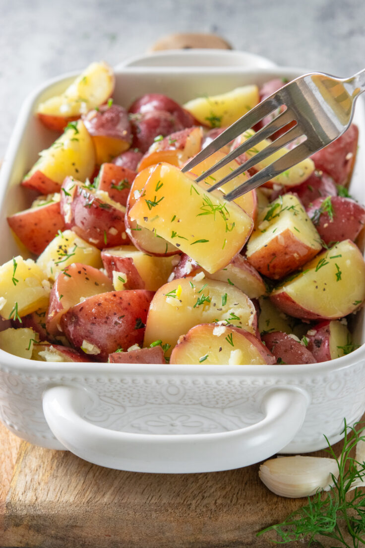A fork lifting a garlic and dill coated red new potato out of the side dish, showing how tender the boiled potato side dish is.