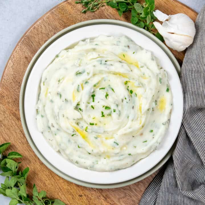 Garlic dill mashed potatoes in a serving dish. You can see melted butter and flecks of fresh green herbs in the potatoes. There are sprigs of fresh thyme, oregano and parsley around the side dish.