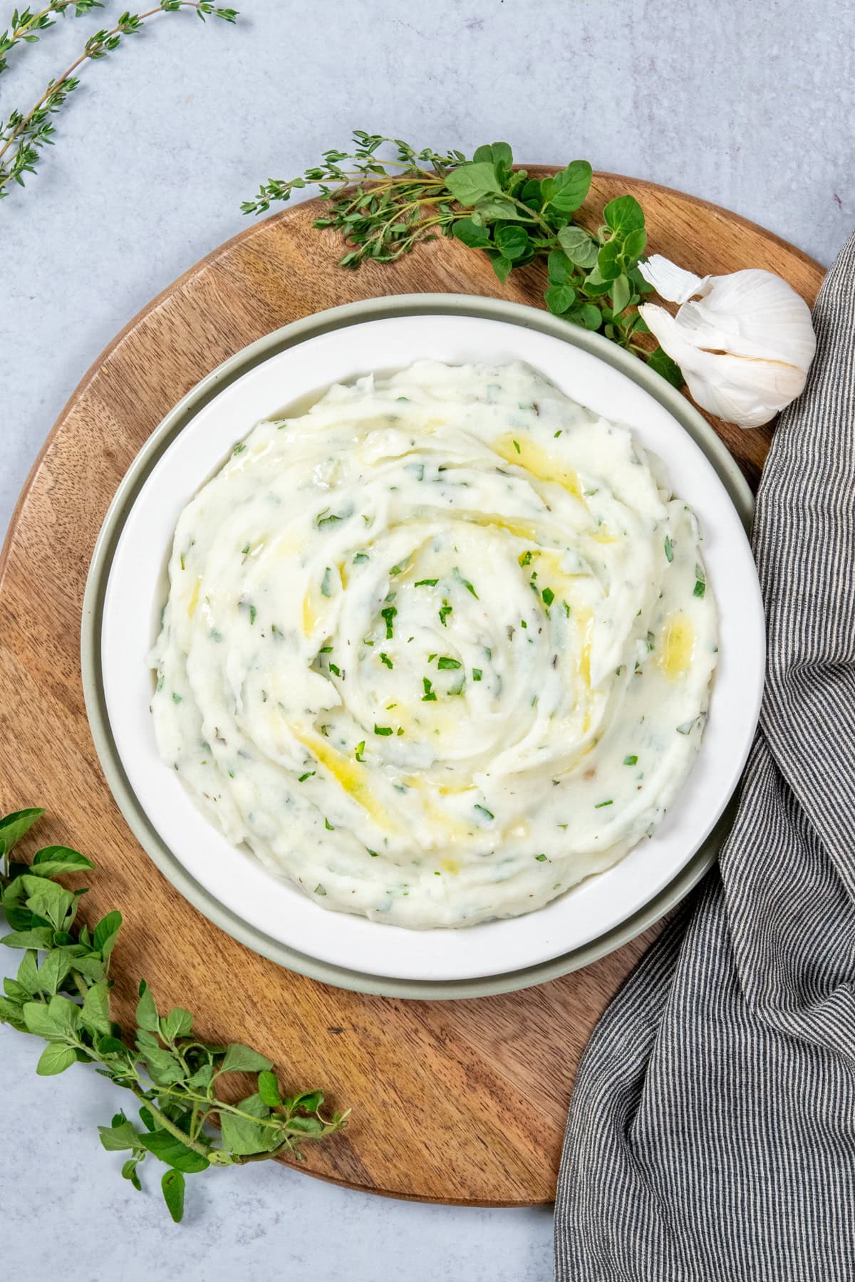 Garlic dill mashed potatoes in a serving dish. You can see melted butter and flecks of fresh green herbs in the potatoes. There are sprigs of fresh thyme, oregano and parsley around the side dish.