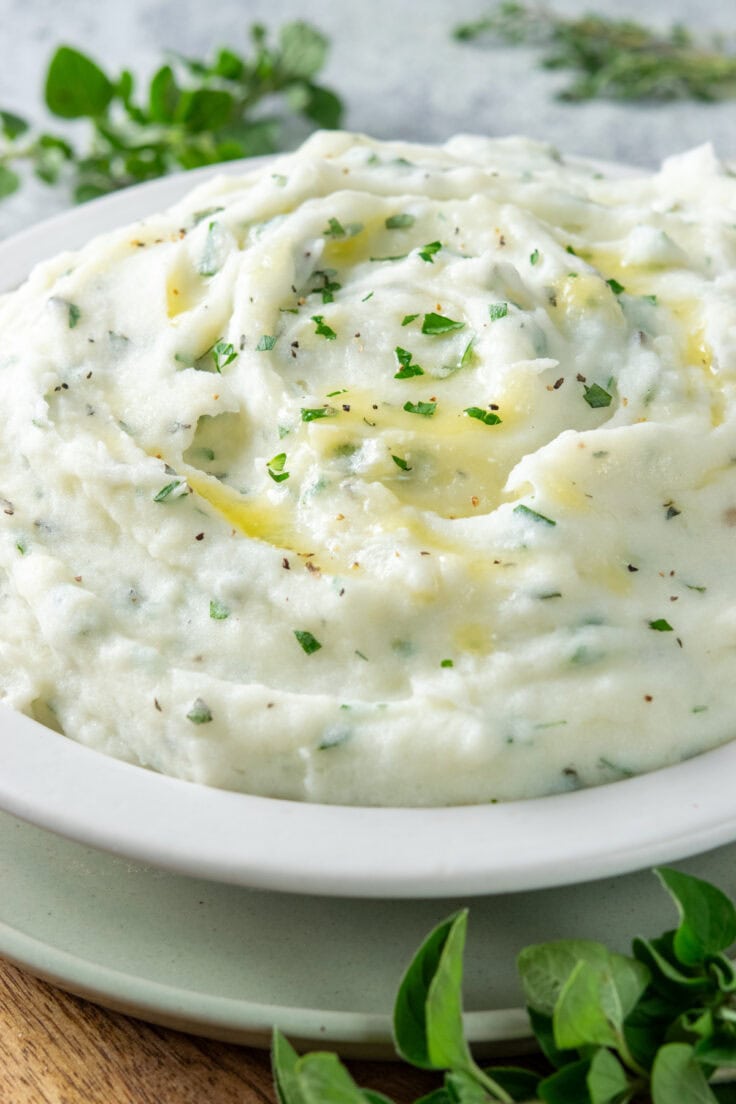A big bowl of freshly whipped garlic herb mashed potatoes. You can see butter on the potatoes and fresh herbs and black pepper. Behind the dish are sprigs of fresh oregano and thyme.