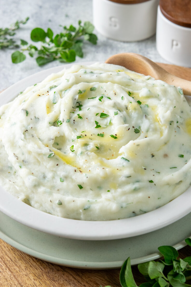 A large bowl of creamy and buttery garlic herb mashed potatoes with a serving spoon in it. Behind the side dish is some fresh roseamary and thyme and salt and pepper pinch pots.