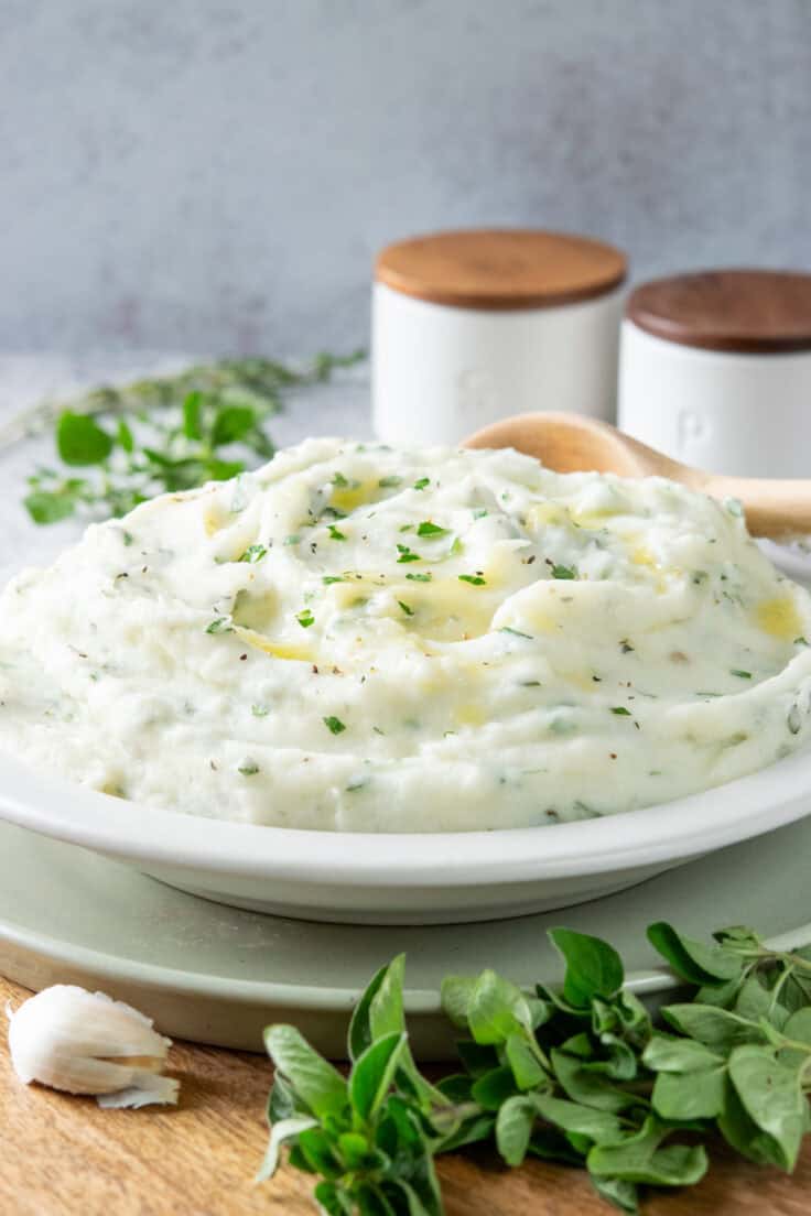 A side dish piled high with the buttery, herb-filled mashed potatoes. In the foreground is a clove of garlic and sprig of fresh oregano.