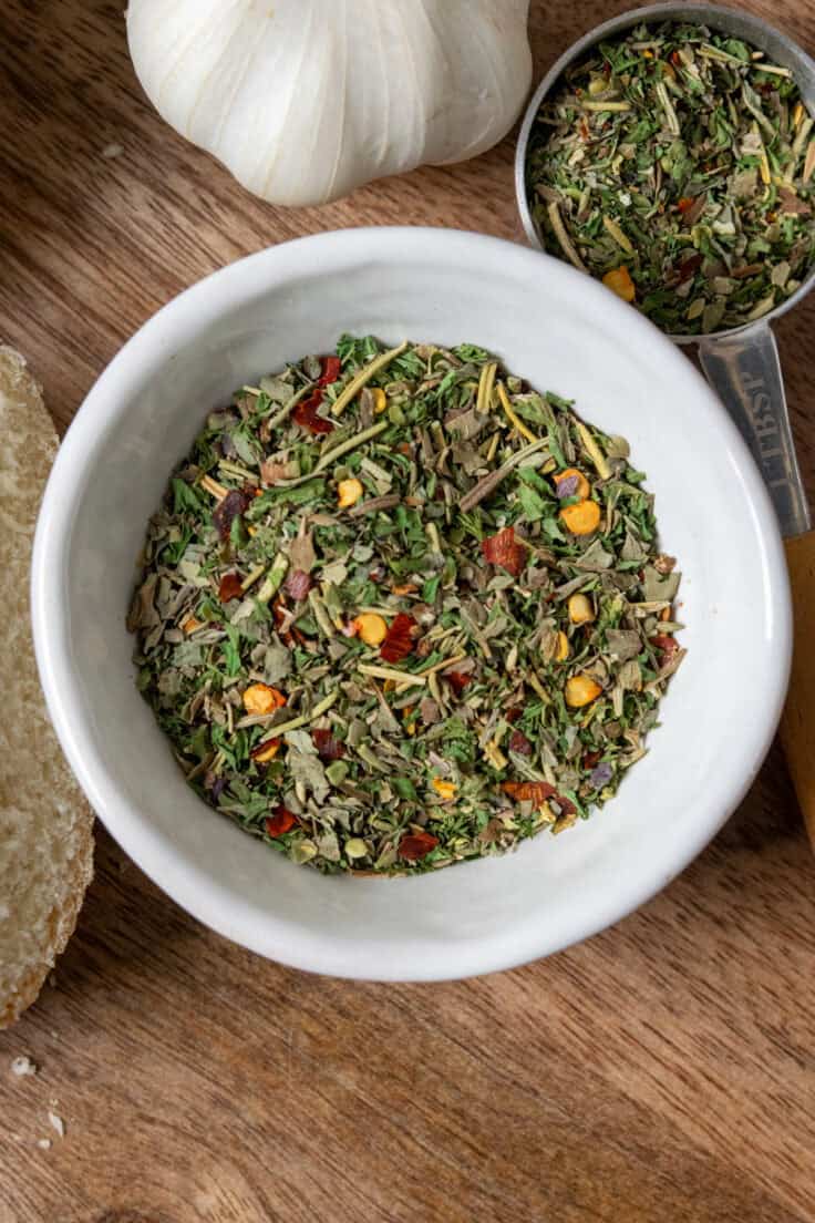A close-up overhead view of a bowl of this Italian seasoning blend for the olive oil dipping sauce for bread. You can see all the colorful herbs and crushed red pepper flakes. Next to the bowl is a tablespoon filled with the spices, ready to combine with olive oil.