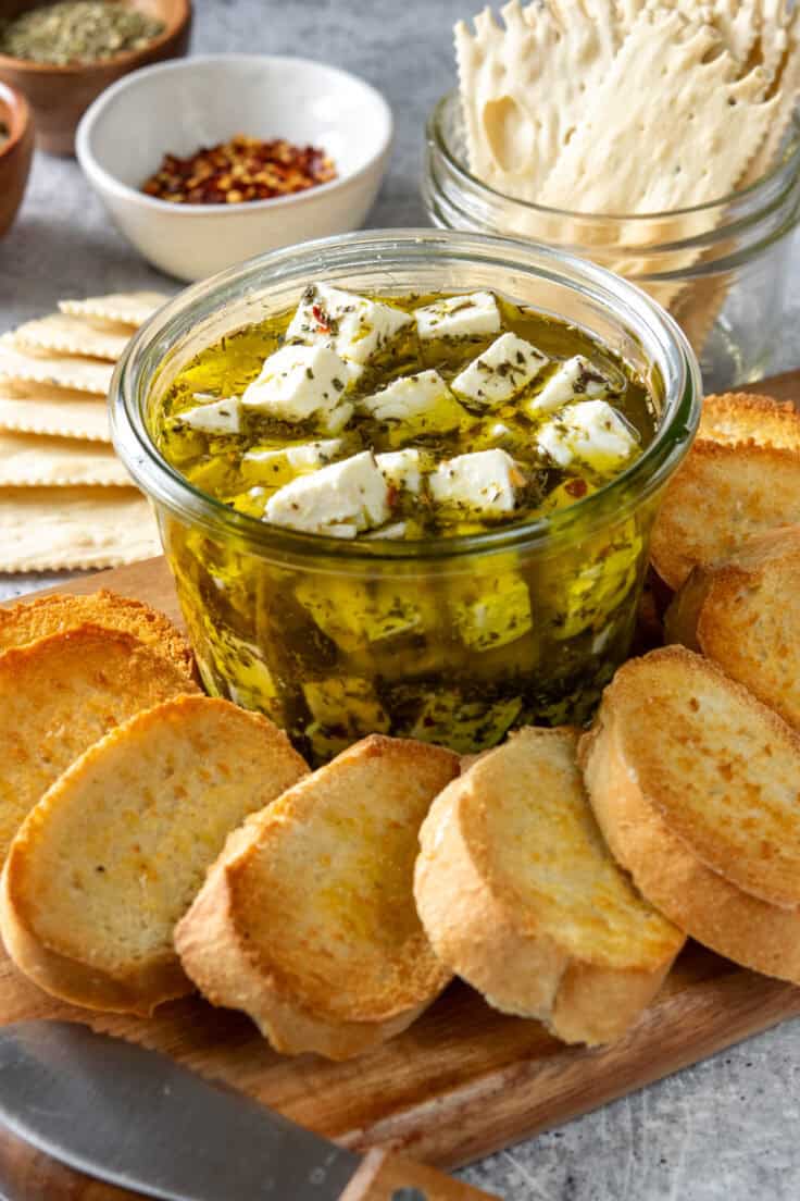 Crostini and crackers on a tray surrounding a bowl of marinated feta.