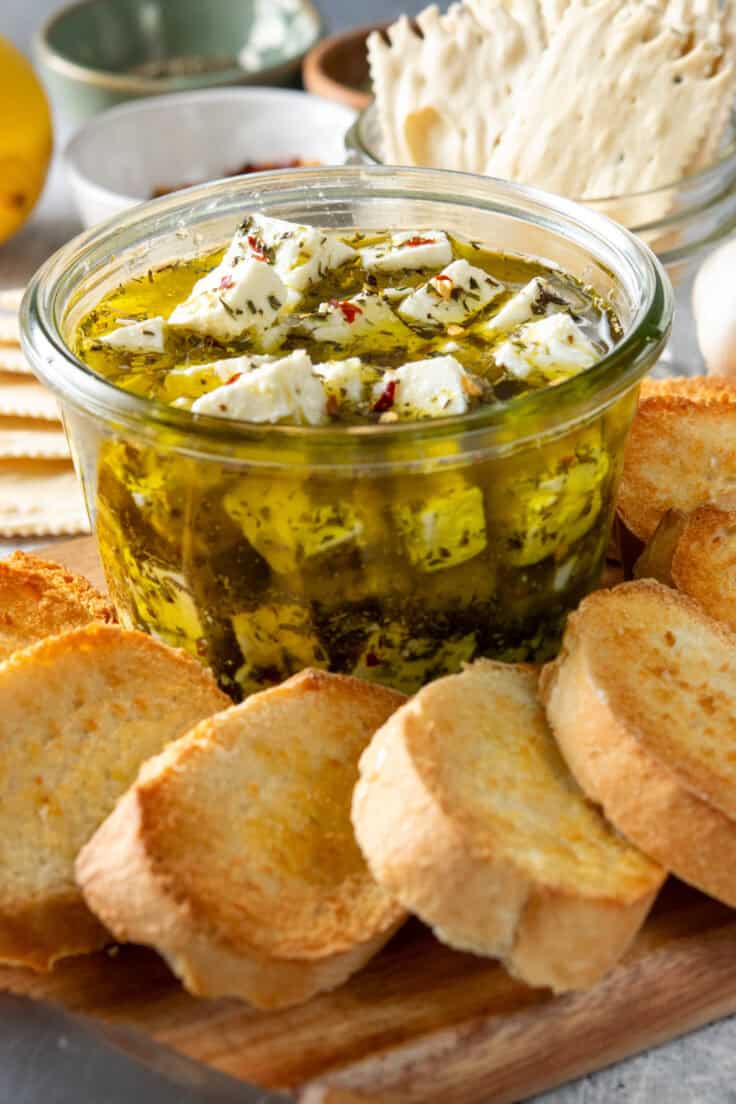 Marinated feta cheese being served with toasted bread and crackers. Behind the feta are small bowls of herbs.