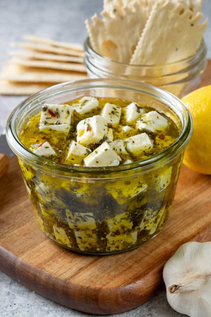 Homemade marinated feta in olive oil with herbs served with crackers. There's a garlic bulb and lemon on the wood tray next to this simple appetizer.