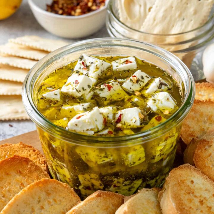 Homemade marinated feta cheese in a bowl with olive oil and herbs, served with crostini and crackers.
