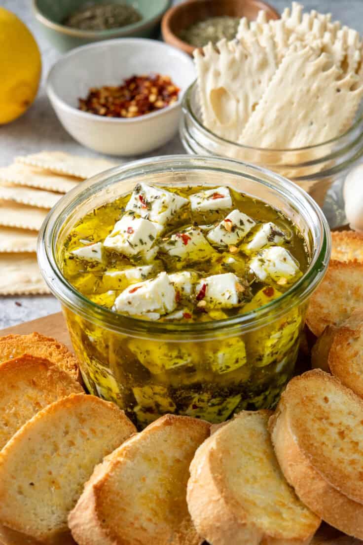 Homemade marinated feta cheese in a bowl with olive oil and herbs, served with crostini and crackers.