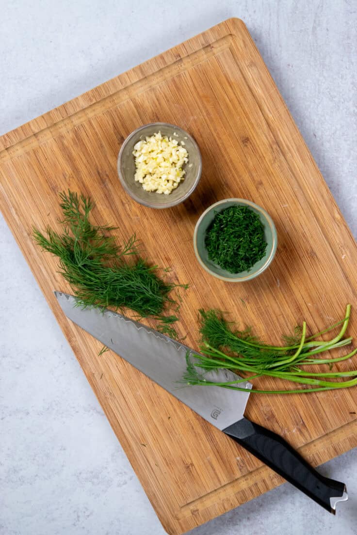 A bunch of fresh baby dill on a cutting board with a knife, showing the thick stems cut off. There's a small bowl with minced dill and another small bowl with minced garlic.