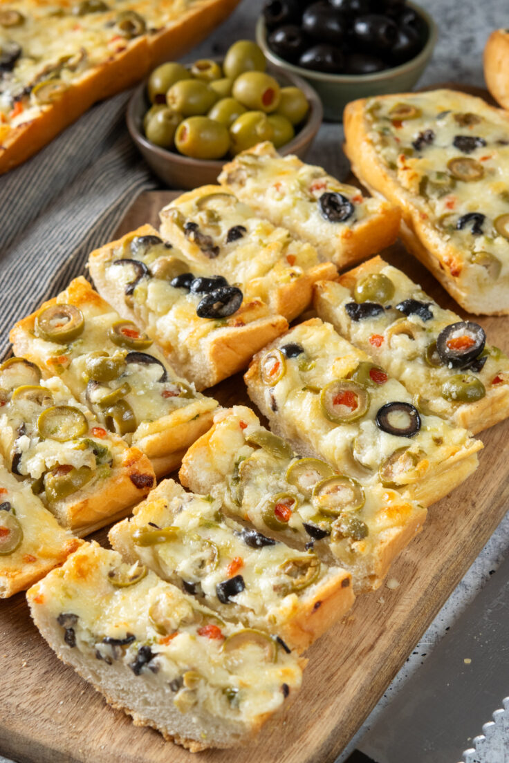 Warm and melty olive cheese bread on a serving board with bowls of green olives and black olives next to the appetizer.