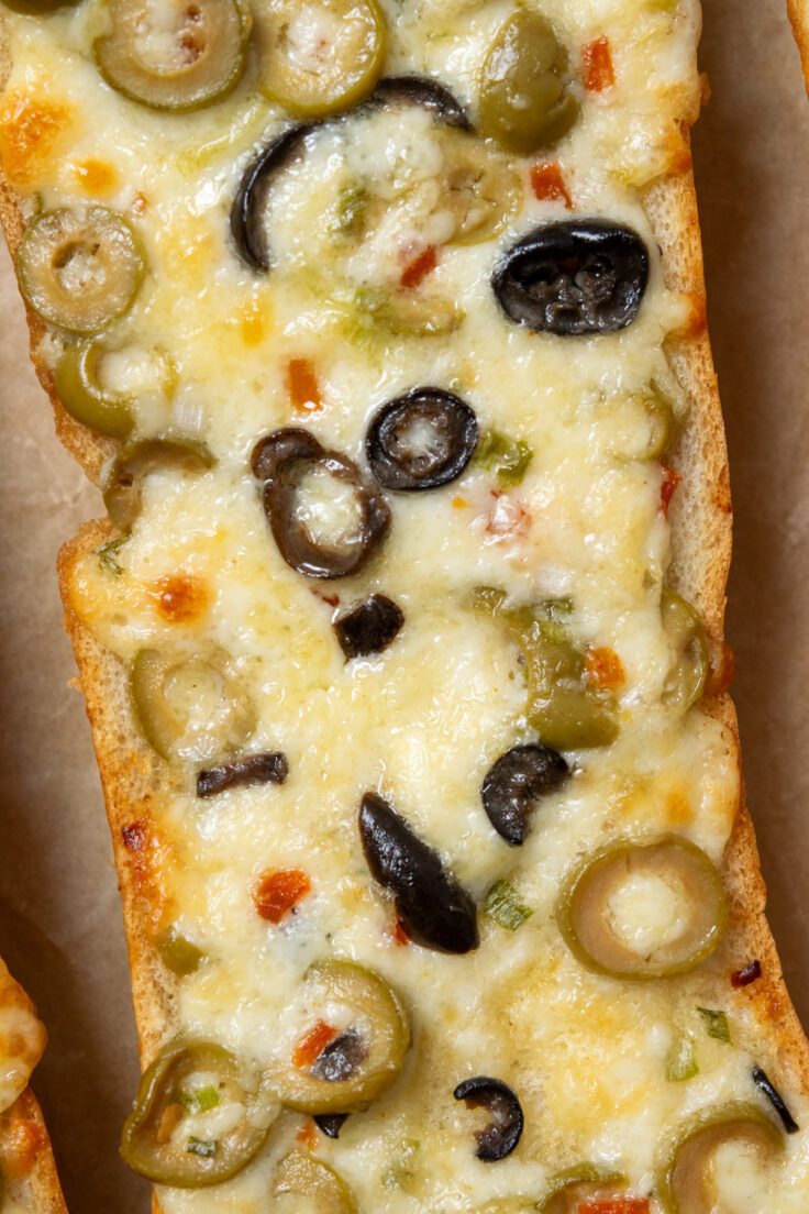 Olive cheese bread just out of the oven. The loaf of bread shows a close up of the gooey melted Monterey Jack cheese with green and black olives melted into the buttery cheesy spread. The bread is crispy on the edges and looks seriously tempting.