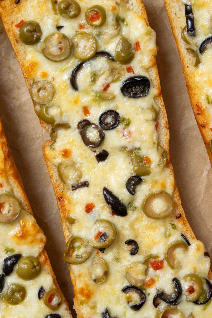 A close up of a hot, melted olive cheese loaf still on the parchment lined baking tray. You can see how buttery and melty the Monterey Jack cheese is with green and black olives and red pimentos melted into it.