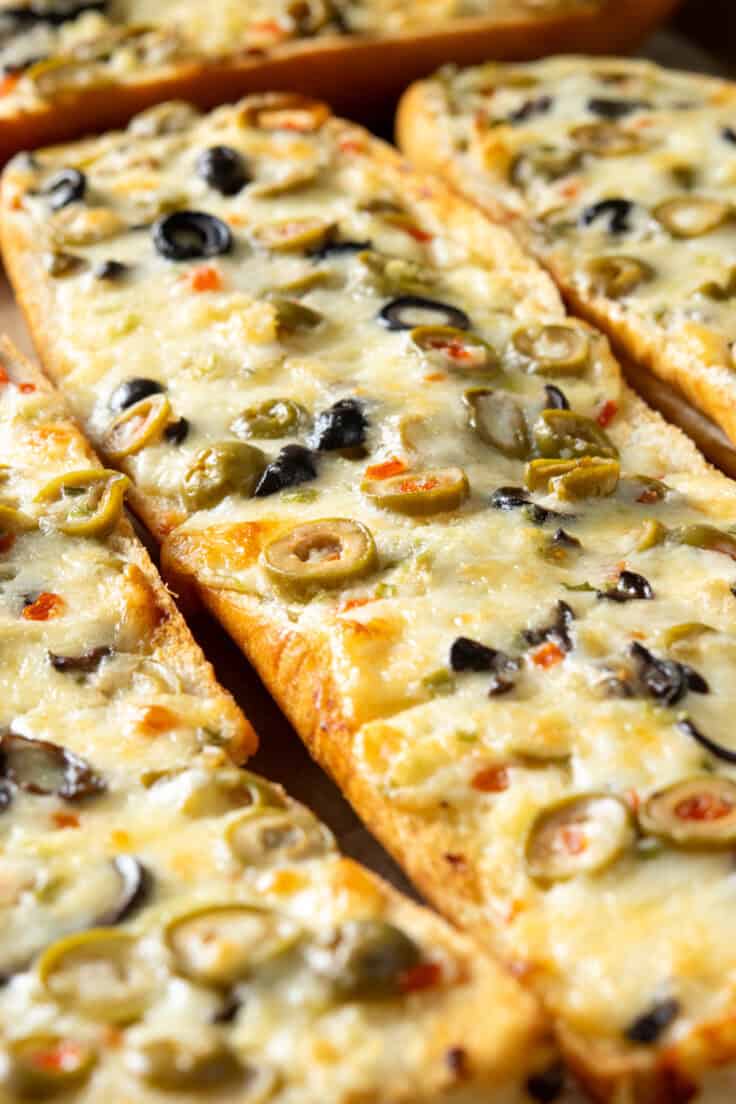 Four halves of olive cheese French bread hot out of the oven and still on the baking tray. You can see the melted cheese that started to brown and a generous amount of black and green olives melted into it.
