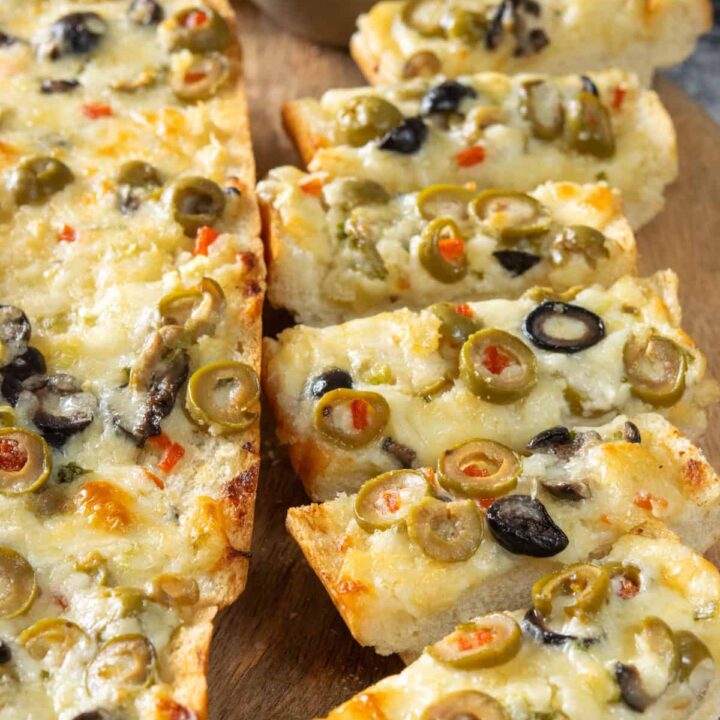 Warm and melty olive cheese bread on a serving board with bowls of green olives and black olives next to the appetizer. One loaf is sliced for an easy finger food and there's a loaf next to it that hasn't been cut yet.
