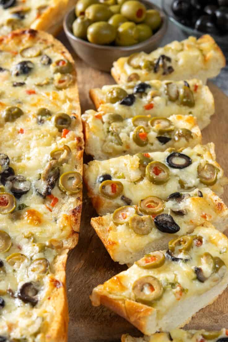 Olive cheese bread cut into thick slices on a serving for an appetizer or party snack. There's an uncut loaf of olive cheese bread and some bowls with green and black olives.