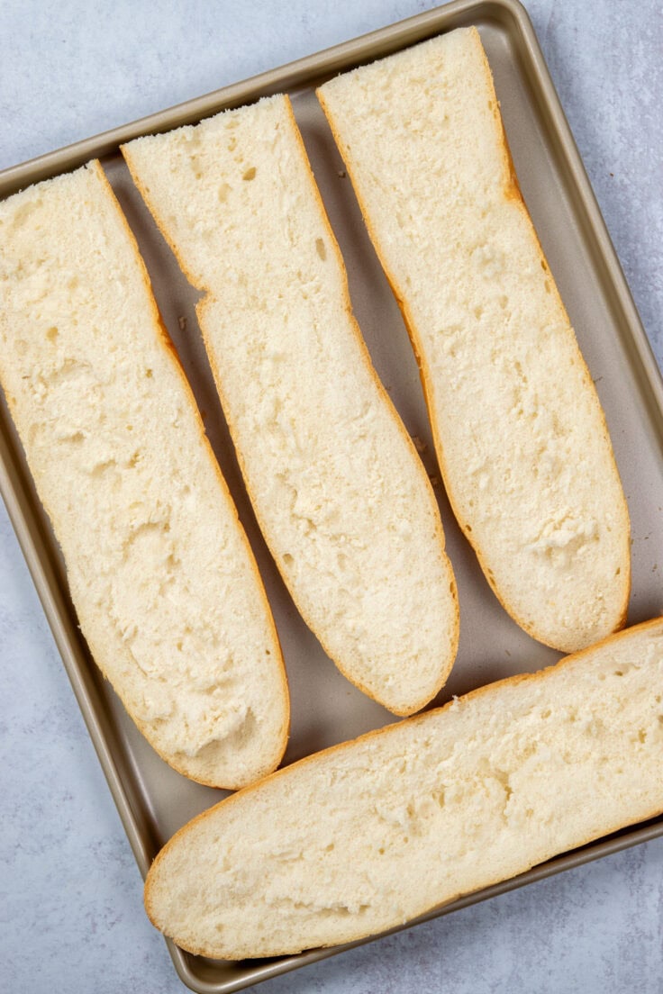 A loaf of French that's been cut into two with each half cut open. The four halves are on a baking tray, bread side up, crust side down.