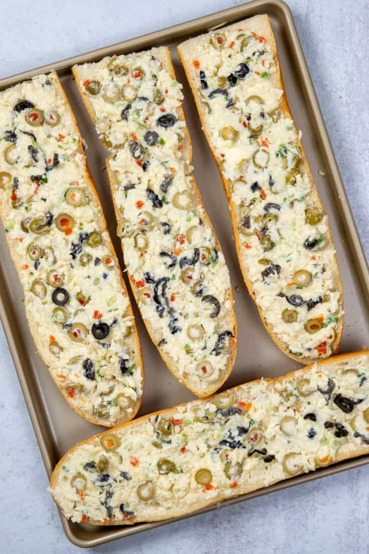 The olive and cheese, butter and mayo mixture spread on the four pieces of French bread on a baking sheet.