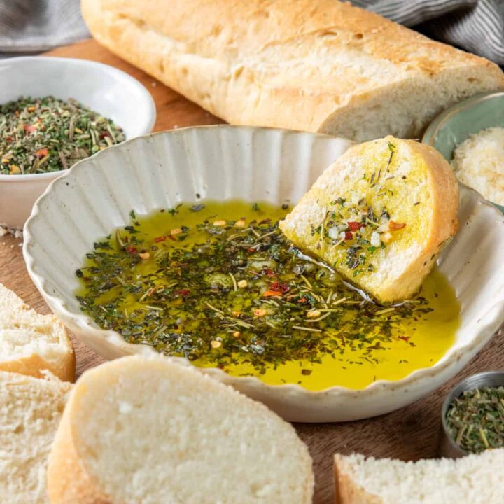 A shallow dish with olive oil dipping sauce for bread. There's a piece of bread dipped in the oil which is loaded with Italian herbs. Next to the dipping sauce is a crusty loaf of bread for dipping.