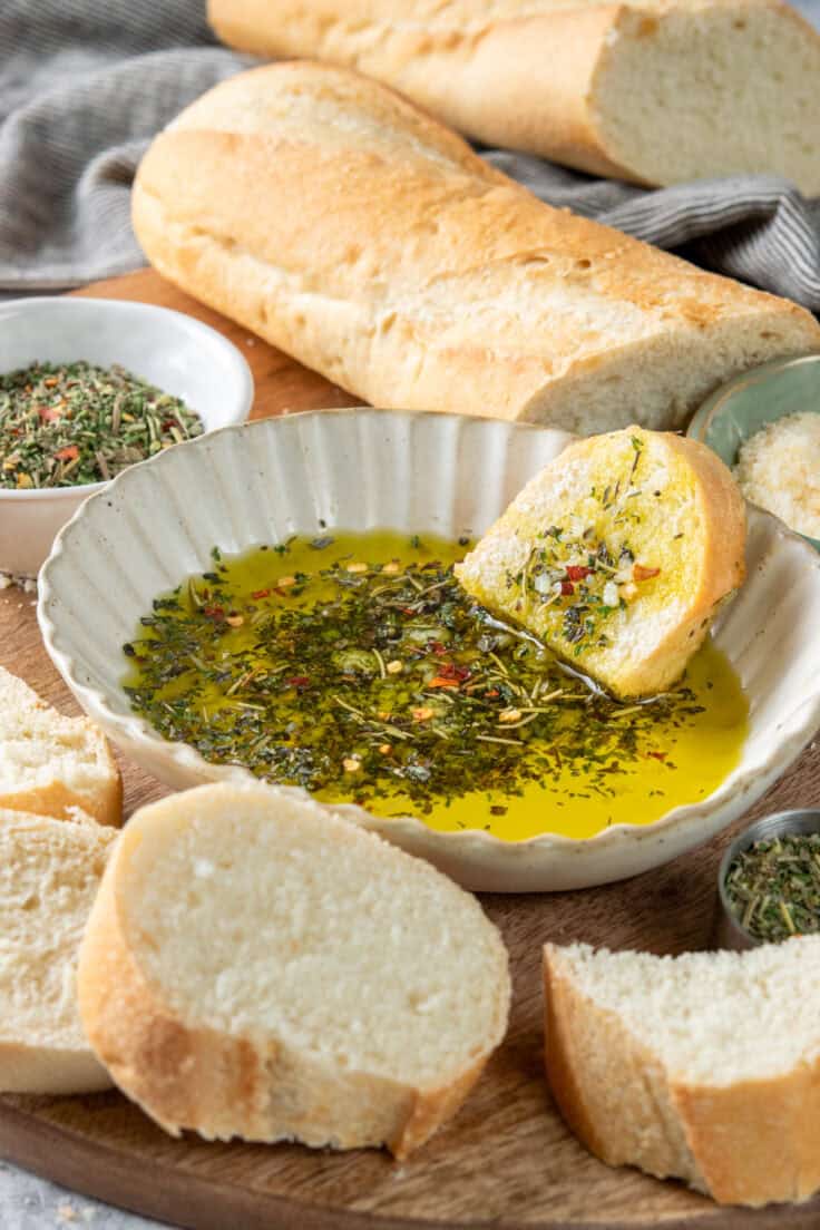 A shallow dish with olive oil dipping sauce for bread. There's a piece of bread dipped in the oil which is loaded with Italian herbs. Next to the dipping sauce is a crusty loaf of bread for dipping.