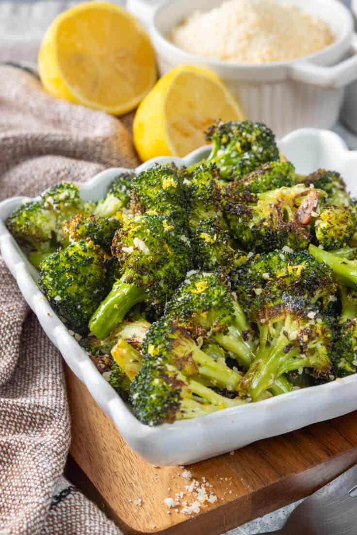 A side dish filled with parmesan roasted broccoli in front of a lemon that's cut in half and a small bowl filled with grated parmesan cheese.
