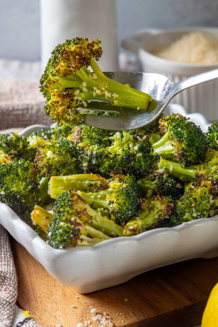 A parmesan broccoli spear on a fork over the side dish. You can see the roasted bits on the florets and bits of grated parmesan.