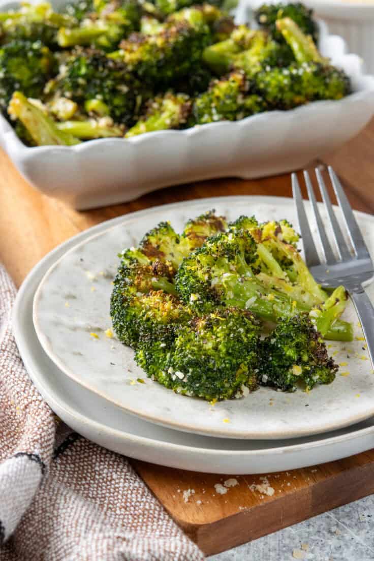Parmesan roasted broccoli on a dinner plate with a fork. You can see some grated parmesan and lemon zest on the roasted broccoli that has caramelized bits on the florets and stems. The plate is in front of the vegetable in a side dish.