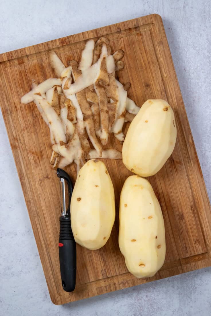 Three russet potatoes on a cutting board being peeled. There is a pile of potato skins next to the peeled potatoes.