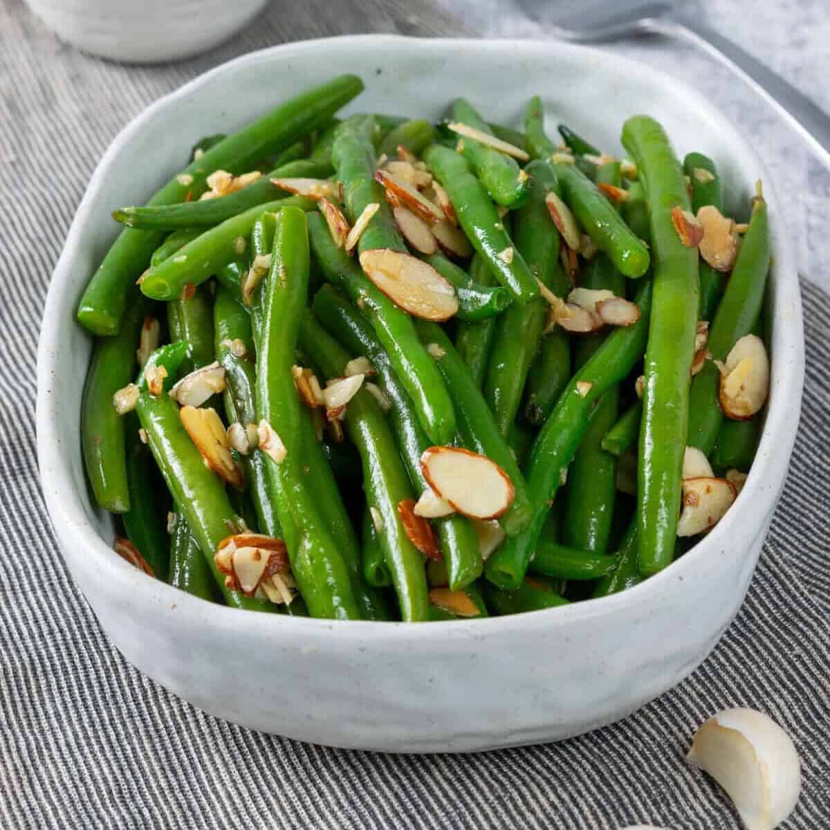 A serving dish with sauteed green beans with garlic and almonds. There's a small bowl of slivered almonds in the background and a few cloves of garlic in the foreground. To the right of the dish is a large serving fork.