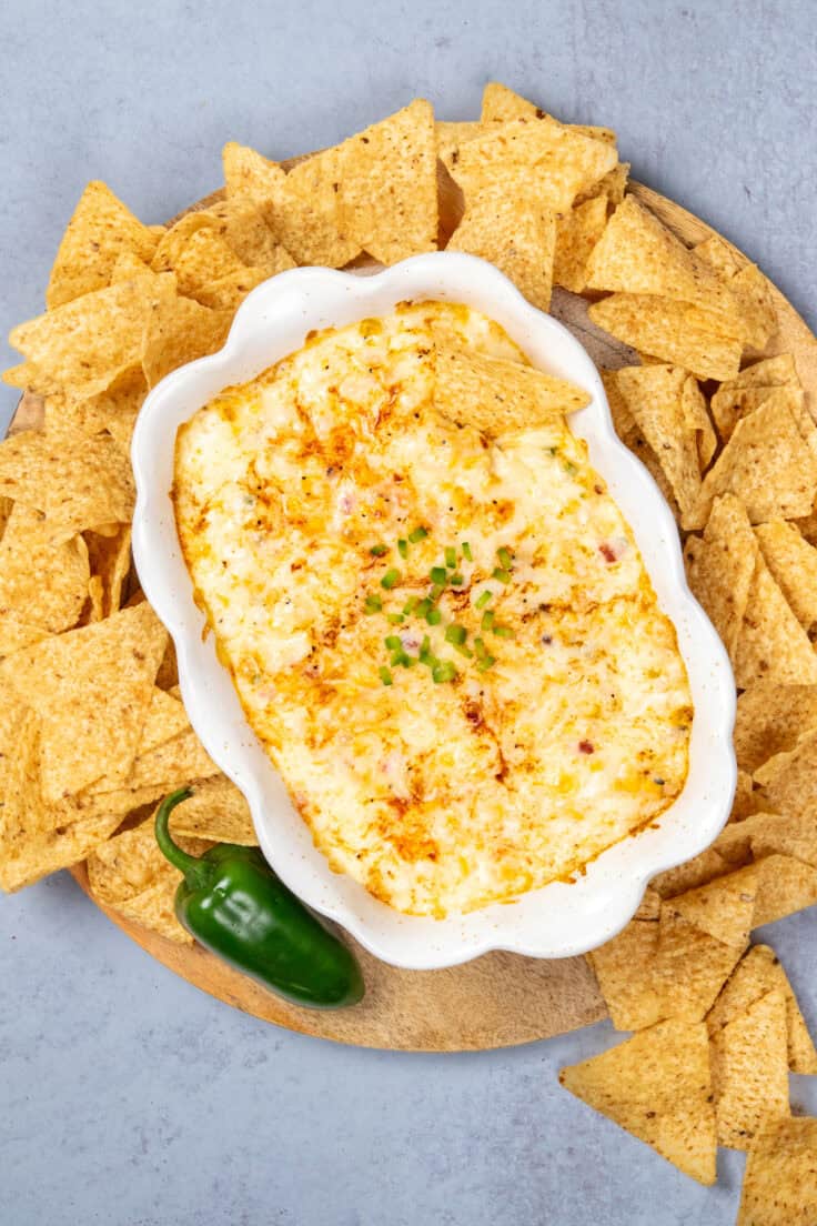 Bubbly hot corn dip covered in melty cheese, in a baking dish on a tray, served with tortilla chips for dipping.
