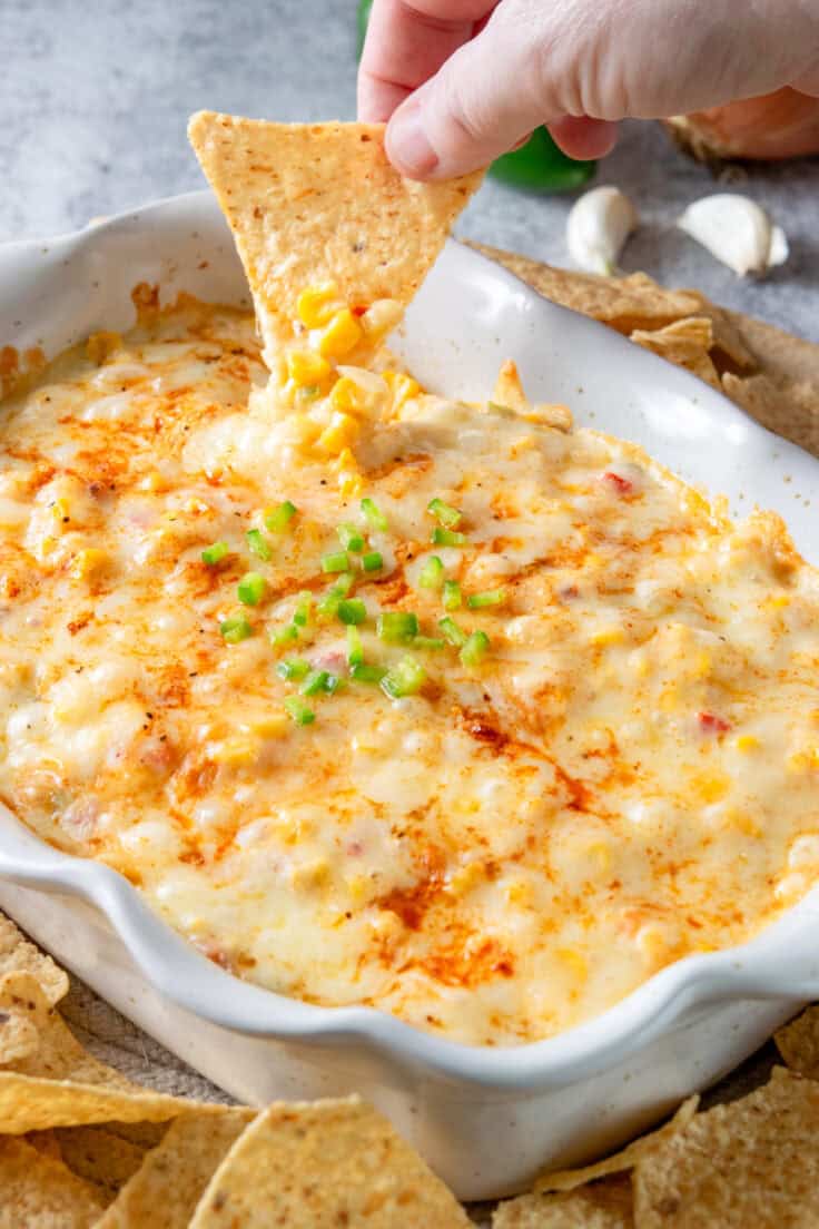 A tortilla chip being dipped into the dip, coming out with a scoop of corn and melted cheese.