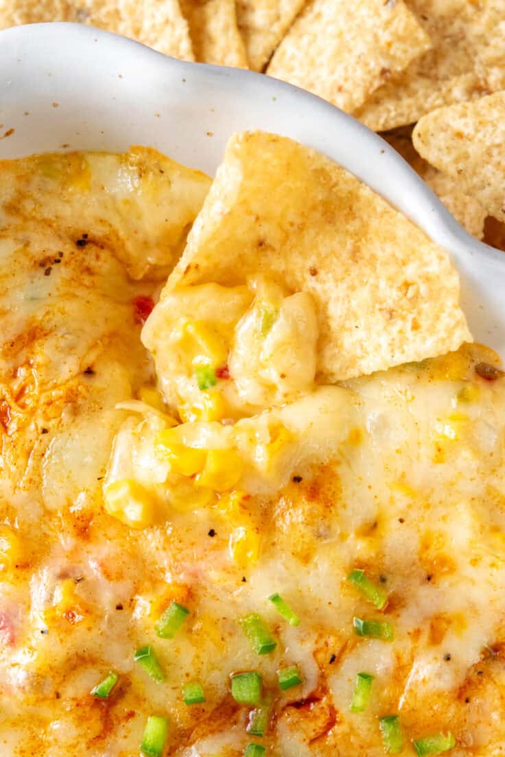 Close-up of a tortilla chip scooping up melted Monterey Jack, corn, and jalapeño from a dish of hot corn dip, showing the gooey cheese, cayenne sprinkle, and tempting texture.