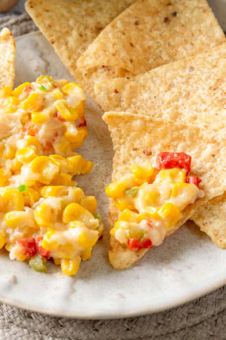 Close-up of a serving of hot corn dip on a small appetizer plate with tortilla chips on the side, including one chip topped with the gooey mix of corn, red peppers, jalapeño, and melted cheese.