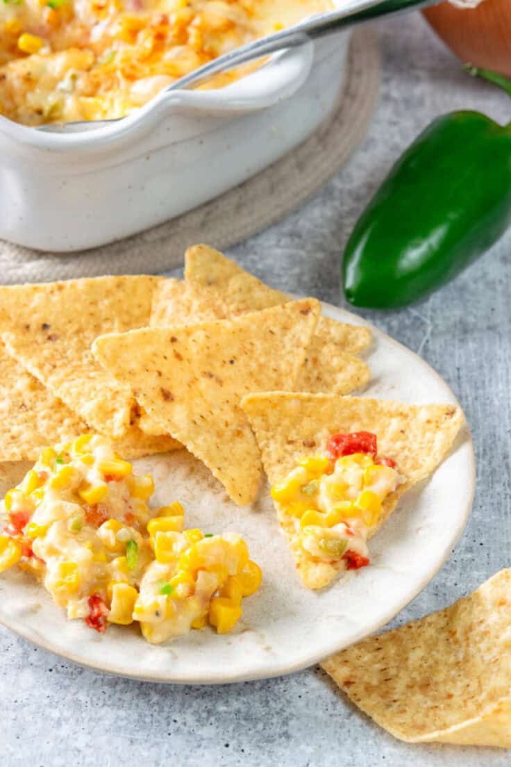Small appetizer plate with a scoop of hot corn dip and tortilla chips next to a baking dish of the dip, with a jalapeño and onion also in the scene.