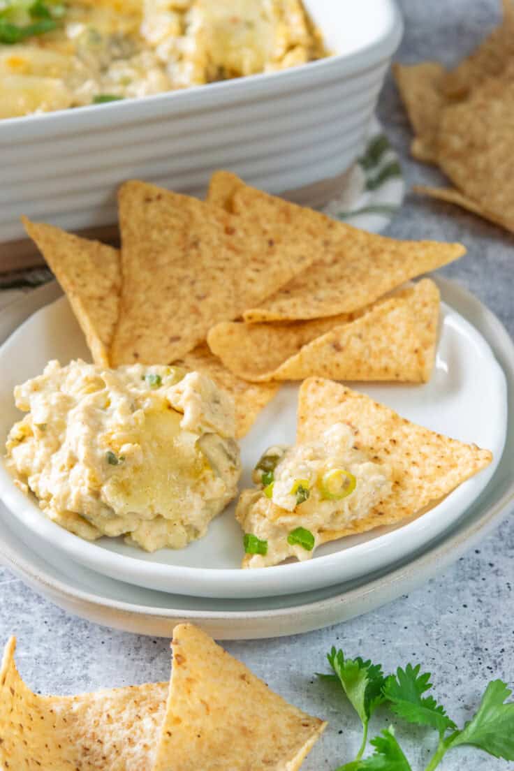 Small appetizer plate with a scoop of warm cheesy enchilada dip and tortilla chips, shown beside the baking dish of dip and a bowl of chips.