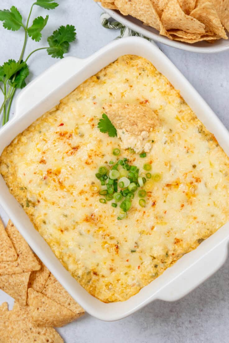 Overhead view of hot chicken enchilada dip in a baking dish topped with green onions, with a tortilla chip dipped in and a bowl of chips on the side.