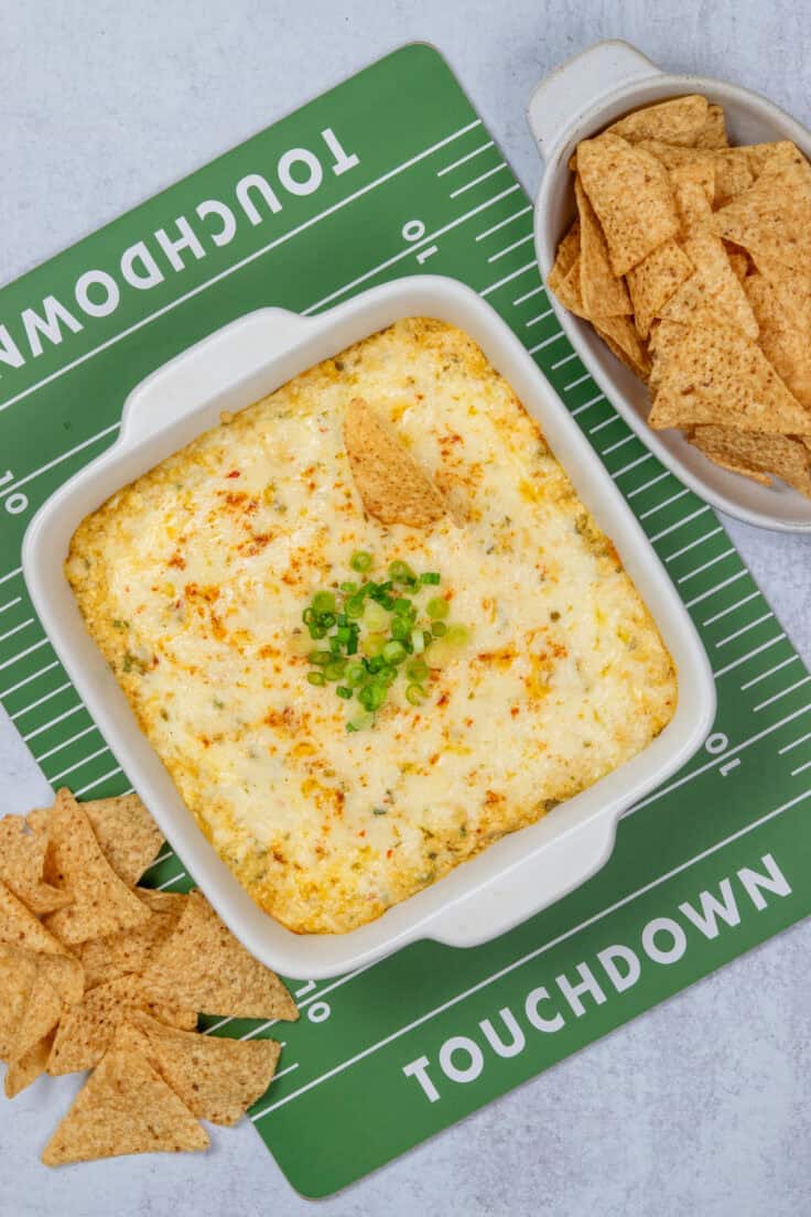 A baking dish filled with bubbly hot chicken enchilada dip, served on a green and white football-themed tray that looks like a football field. There's a bowl of tortilla chips next to the party dip that's perfect for football Sunday, the Superbowl, Christmas parties, Cinco de Mayo and more.