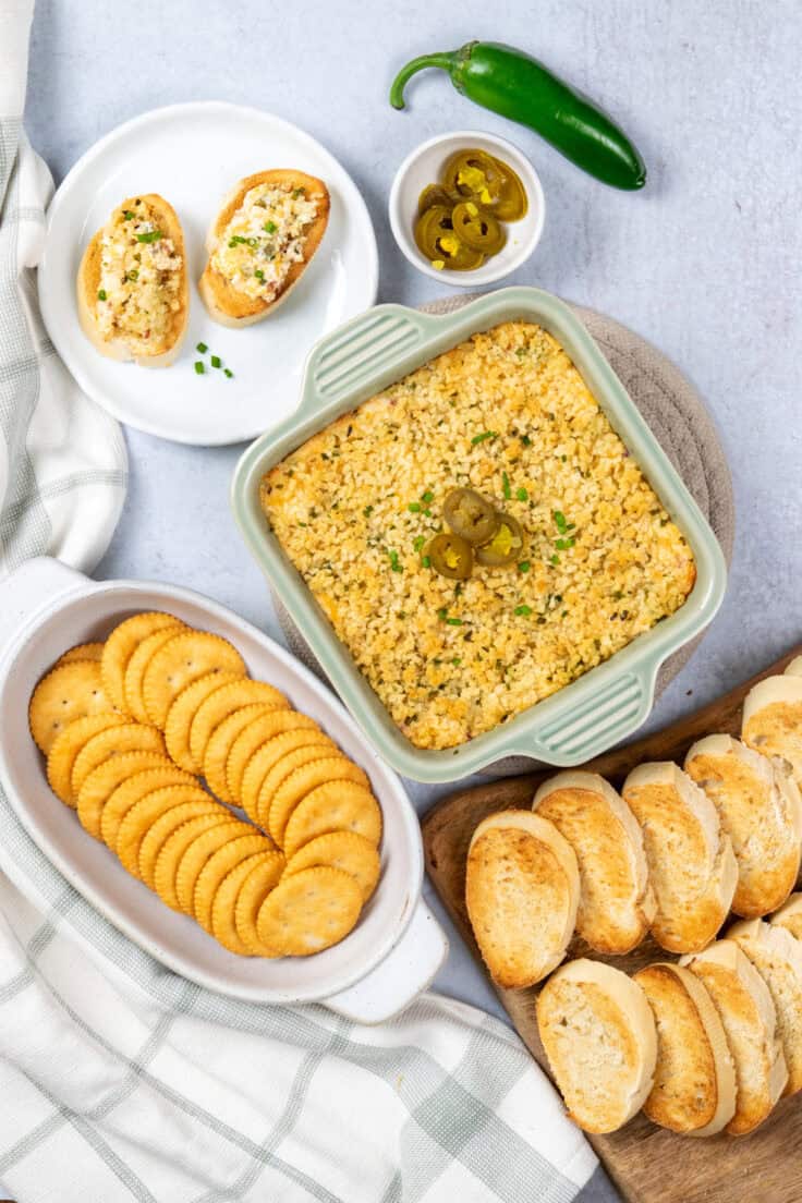 Overhead view of baked jalapeño popper dip with crackers, sliced French bread, and mini appetizers ready for serving.