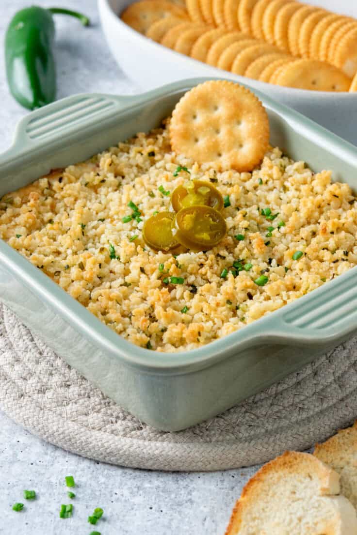 Close-up of cheesy jalapeño popper dip with pickled jalapeño slices and a cracker being dipped in.